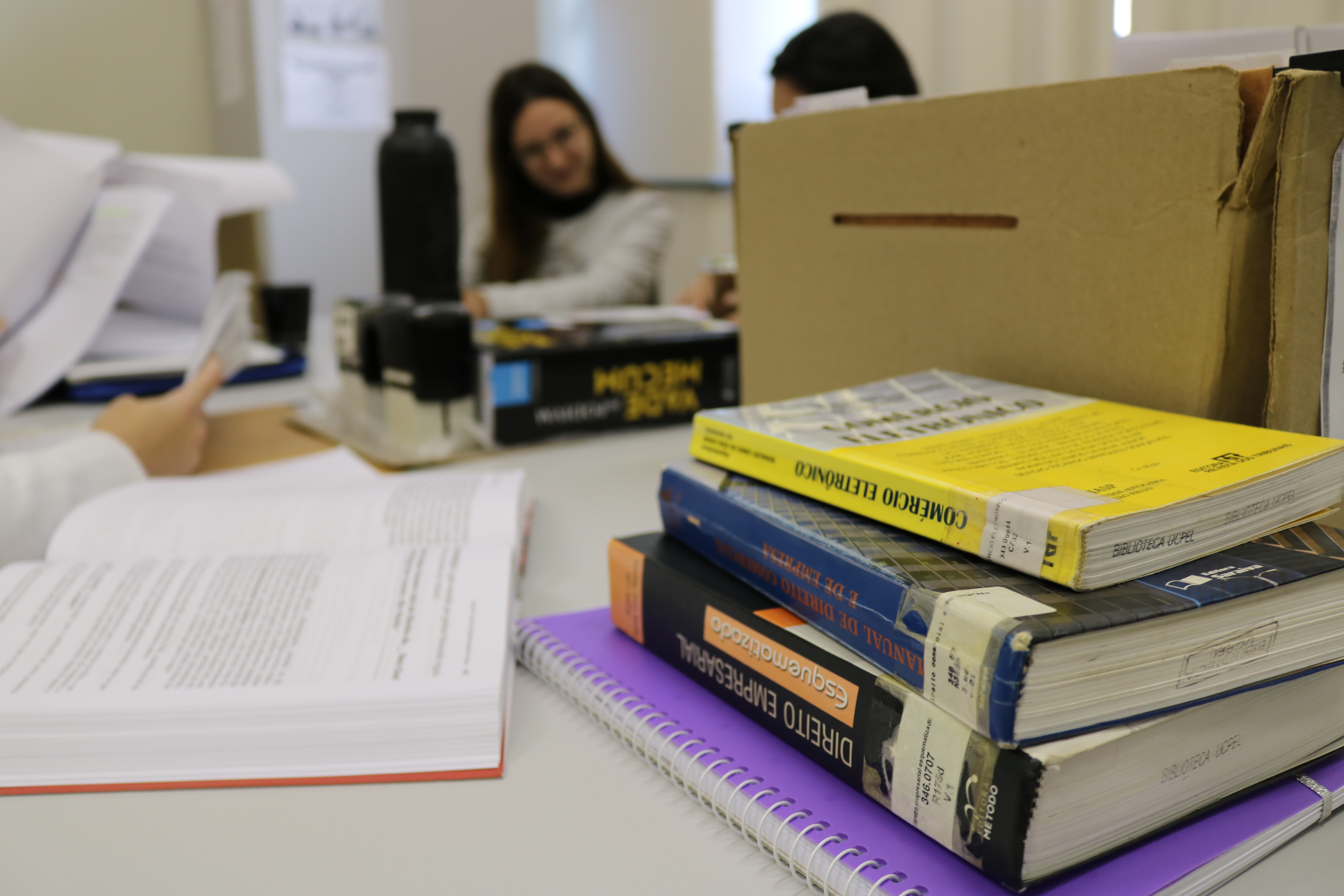 livros sobre direito empilhados em uma mesa com acadêmicas do curso conversando ao fundo