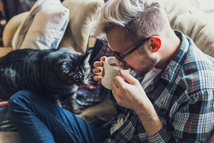 homem loiro e com óculos tomando café, sentado em um sofá com seu gato ao lado