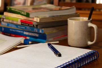 caderno aberto em cima de uma mesa com uma caneta em cima, uma xícara com colher ao lado e outros livros à frente
